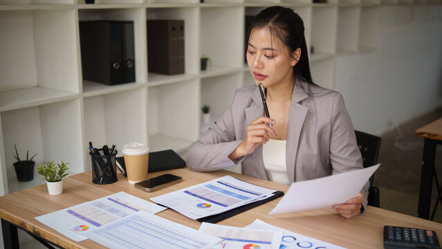 Thoughtful Asian businesswoman evaluating printed financial reports at her desk.