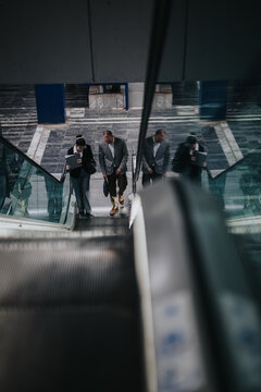 Business people ride an escalator in a modern office lobby carrying bags and documents. A top-down view captures professionals commuting together in a glass-and-steel atrium.