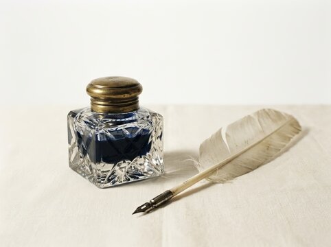 Vintage glass inkwell filled with blue ink next to a feather quill pen on a white linen surface