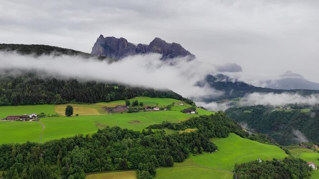 Jagged peaks emerge above low clouds, with green meadows below in Dolomites, South Tyrol, Italy. Drone footage