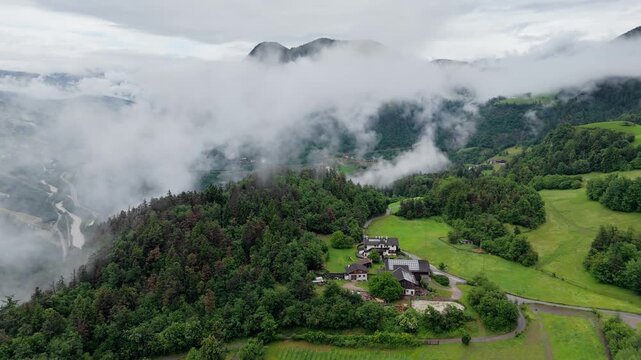 Alpine farm beside green meadows and forest, with low clouds over mountain valley in Dolomites, South Tyrol, Italy