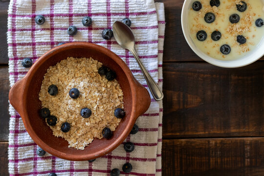 Horizontal aerial view of a bowl of oatmeal and blueberries on a kitchen tablecloth and a rustic wooden table next to a small bowl of yogurt and blueberries, depicting a healthy breakfast or snack