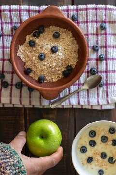 A vertical aerial view of a hand holding a green apple with water droplets next to small bowls of healthy food on a rustic wooden table