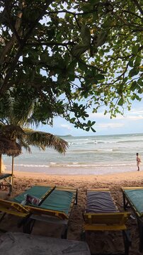 Sun loungers on sandy beach with ocean waves under shade of tropical trees in Sri Lanka