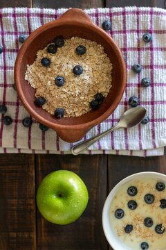 A vertical aerial view of a green apple with water droplets next to a bowl of oatmeal with blueberries and another bowl of yogurt on a rustic wooden table for a healthy breakfast