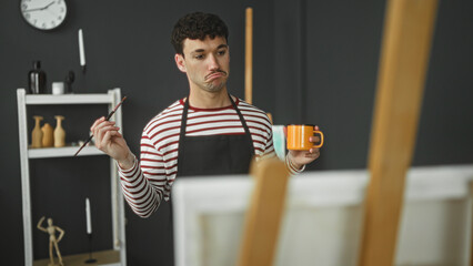 Man painter wearing apron and striped shirt holds paintbrush in hand and drinks from orange mug while looking at canvas in studio  thoughtful pause. © Krakenimages.com