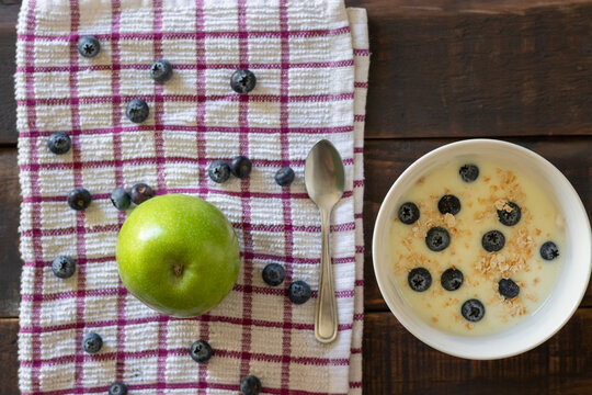 A bird's-eye view of a healthy breakfast or snack consisting of a bowl of yogurt with blueberries and a green apple on a rustic wooden table