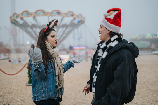 A young couple wearing Christmas accessories talk animatedly outdoors. The pair stand near blurred amusement rides at a festive winter fair.