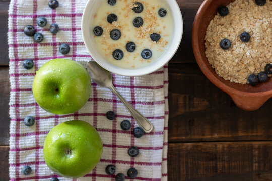 A bird's-eye view of two freshly washed green apples on a tablecloth next to a small bowl of yogurt and another with oatmeal and blueberries, ready for breakfast or a healthy snack