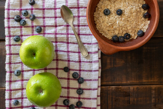 A bird's-eye view of two freshly washed green apples on a tablecloth next to a small bowl of oatmeal with blueberries, ready for breakfast or a healthy snack