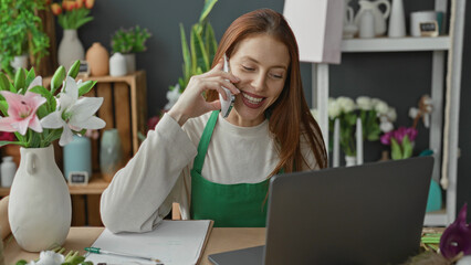 Obraz premium Woman florist holding smartphone to ear with hand on laptop and arranging flowers in a building; cheerful small business.