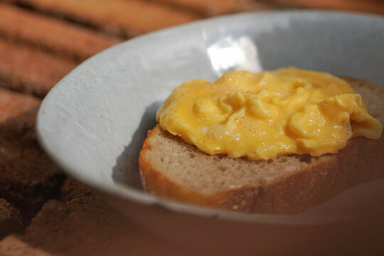 Delicious scrambled eggs on a slice of toasted sourdough bread served in a rustic ceramic bowl under warm natural sunlight.