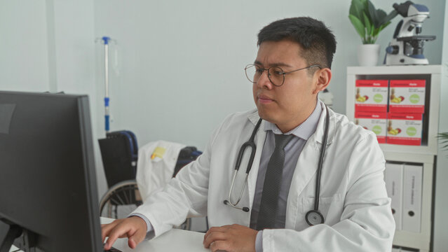 Man doctor at computer typing on keyboard while squinting with closed eyes and stethoscope visible at desk in building; medical fatigue.