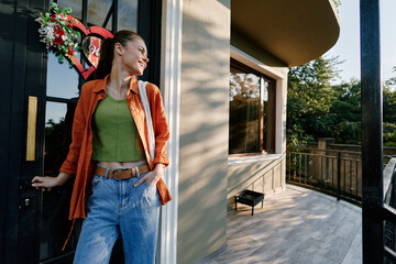 Young woman in orange shirt and green top opens the front door of her house. Smiling girl in jeans stands on the porch near the entrance. Home and lifestyle concept. © SHOTPRIME STUDIO