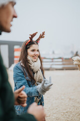 A teenage girl wearing a reindeer headband and scarf smiles while interacting with others. She enjoys a casual outdoor winter holiday gathering with friends and family members nearby. © qunica.com