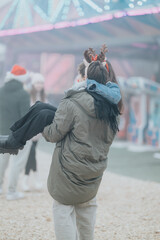 Naklejka premium A young couple at a holiday fair with the man carrying his girlfriend as she wears reindeer antlers. They embrace under festive lights at an outdoor winter amusement area.