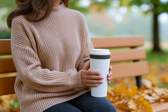Woman in beige sweater holding a white coffee cup while sitting on a wooden bench surrounded by autumn leaves in a park setting