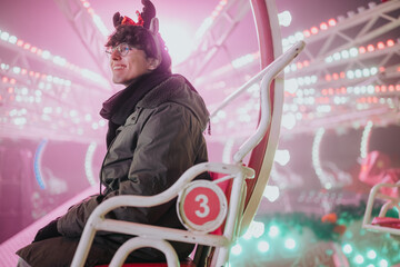 Naklejka premium A young man wearing reindeer antlers and glasses smiles while seated on a lit carnival ride. The festive scene shows colorful lights and joyful atmosphere with friends at night.