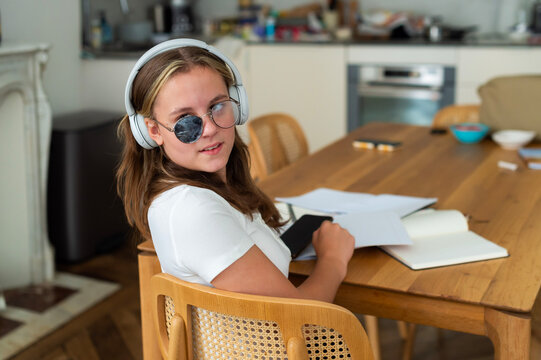 A schoolgirl in Paris, France, turns toward the camera while wearing strabismus correction glasses with an eye patch. She sits at a wooden table with study materials and headphones.