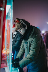 Fototapeta premium A young man in a parka and reindeer headband plays a claw crane game at a festive night market. The candid scene shows friends enjoying a neon-lit arcade booth during a cold holiday evening.