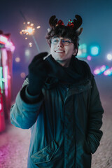 A young man wearing reindeer antlers and glasses holds a lit sparkler in hand. He enjoys a neon-lit winter night scene with friends at an outdoor market. © qunica.com
