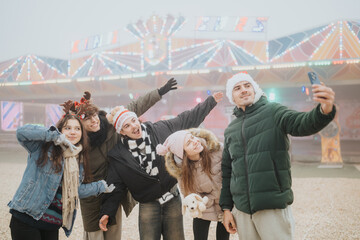 Naklejka premium A group of young friends wearing Santa hats and winter coats take a selfie together. They pose happily in front of colorful carnival lights at an outdoor holiday fair.