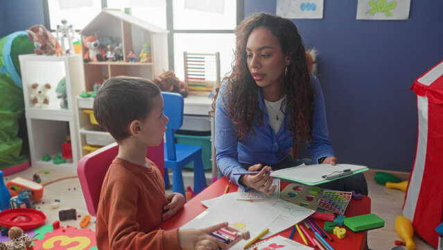 Boy student draws with crayon while teacher holds a clipboard and points pen in preschool building; calm concentration.