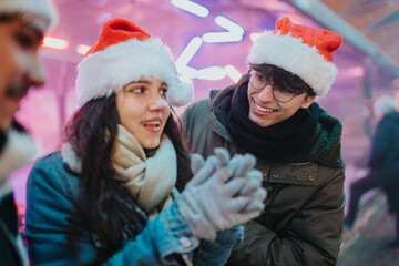 Naklejka premium A young couple of friends wearing Santa hats smile and chat at a festive outdoor market. The cheerful pair share a warm moment at a winter holiday market with colorful lights.
