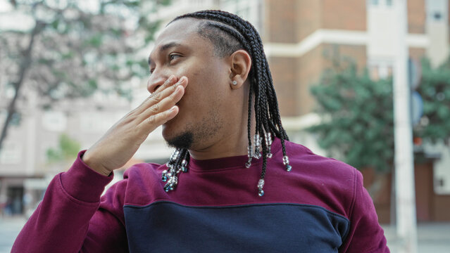 Young man with braided hair and beads holds hand to mouth gesture on a street; thoughtful reflection.