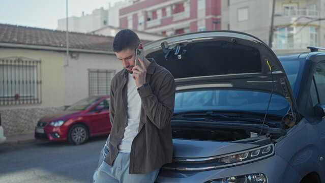 Man holding phone to ear beside car with open hood on street, leaning on bumper and looking up; roadside vehicle trouble concern.