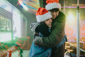Naklejka premium Young couple wearing Santa hats embrace warmly while smiling at a festive outdoor market. A cozy holiday hug captures seasonal joy and togetherness amid colorful lights.