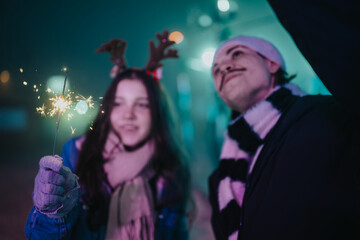 Naklejka premium Two friends hold a lit sparkler while enjoying a festive winter evening. A cozy holiday moment with warm accessories and soft lights in the background.