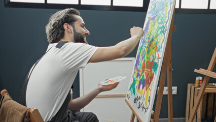 Young hispanic painter man painting on canvas with palette and brush, arm extended, wearing apron, seated at easel with colorful abstract brushstrokes in studio  creative concentration inspiration. © Krakenimages.com