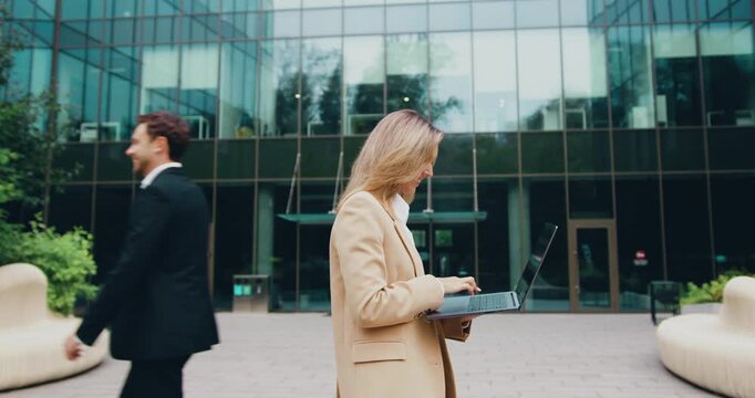 Side view of a smiling young woman with long hair using her laptop, typing a message while walking along a street toward an office building next to green bushes outdoors.