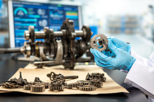 Gloved technician holds a metal gear beside a disassembled engine shaft and parts in a modern workshop, with a diagnostic screen in the background.