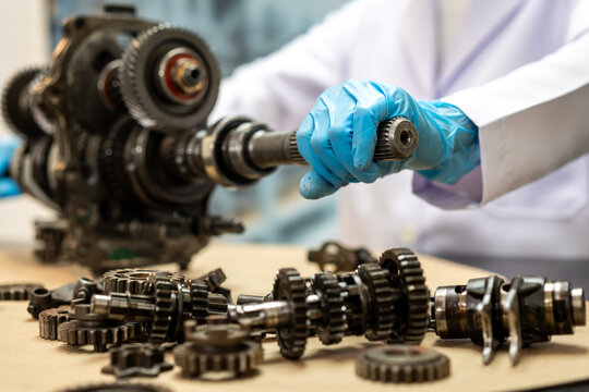 Mechanic in blue gloves holds a metal shaft beside a disassembled gearbox, with gears and machine parts spread across a workshop table.