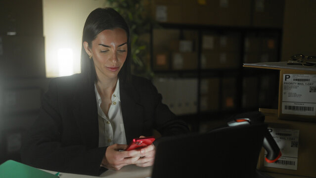 Woman holding a red smartphone and typing on a laptop, smiling while working in a dim storage building; night shift contentment.