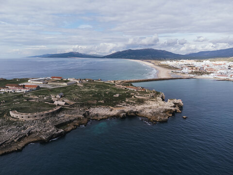 Aerial view of Isla de las Palomas, a historic coastal fortification on a rocky peninsula overlooking a bay with white town and sandy beach beneath mountain range in Strait of Gibraltar, Spain
