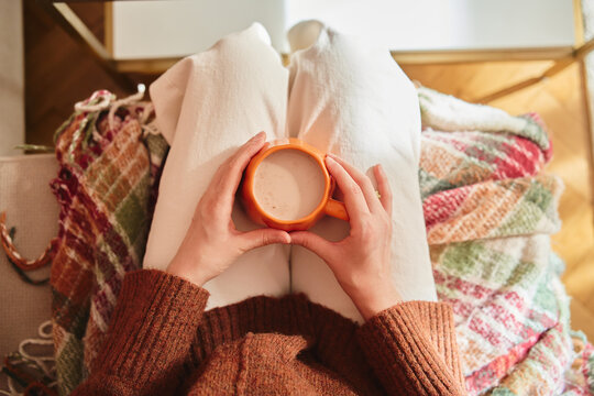 Top view of a woman in a brown sweater sitting on a sofa with a cozy autumn blanket, holding a warm drink in an orange mug on her lap. Cozy home atmosphere, fall aesthetic,soft natural light.