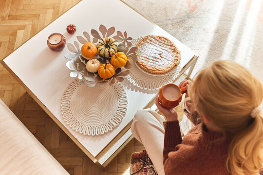 Top view of a young woman with a cup of hot drink at a cozy table with pumpkin pie, decorative mini pumpkins, and autumn candles. Cozy home, fall aesthetic, hygge, warm seasonal atmosphere.