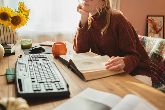 Woman studying at a cozy wooden desk with open book, warm drink, and sunflowers in a softly lit home interior.