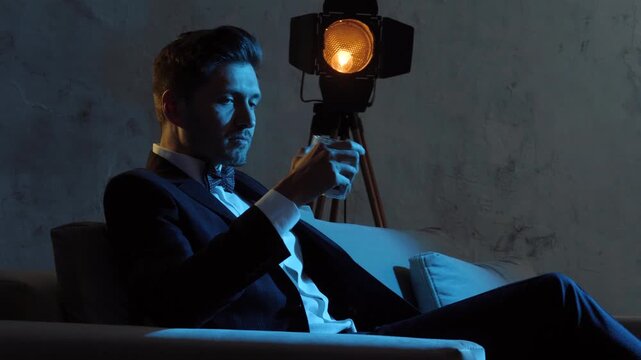 Man in dark suit seated on sofa lifting glass to lips under warm studio spotlight with cool blue rim light in lounge interior