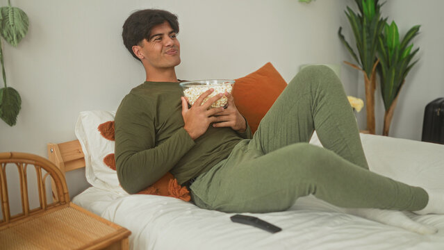 Man on bed holding popcorn bowl, hand to mouth eating and smiling in a building bedroom; cozy relaxed leisure time.