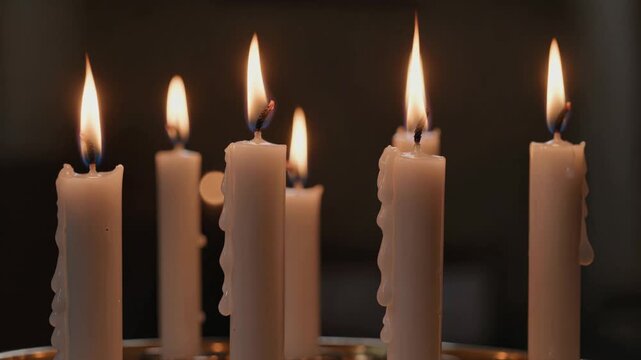 Cinematic video of several white candles burning with warm flickering flames and melted wax against a dark softly blurred background.