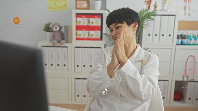 Woman doctor in white coat with yellow stethoscope raises fists and claps hands in a pediatric clinic building; telemedicine success joy.