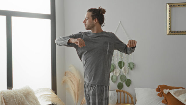 Man stretching arms beside bed in a sunlit bedroom in a residential building, reaching toward window while wearing pajamas and looking sideways; calm morning.