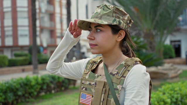 Woman soldier salutes with right hand wearing camouflage vest and visible us flag patch in street; pride duty.
