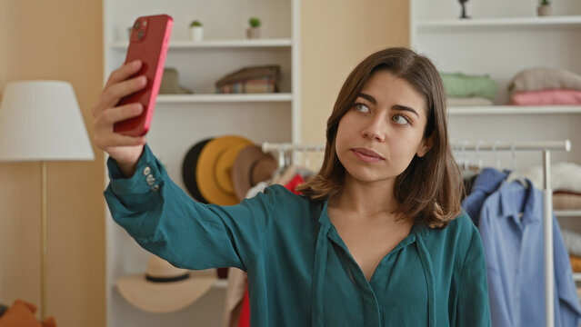Woman holds pink smartphone at arm's length taking a selfie with exposed collarbone in a clothing studio; contemplation.