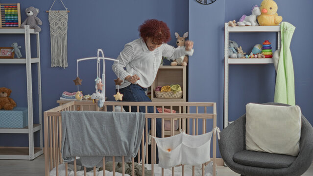 Woman jumps with arms raised beside wooden crib with hanging mobile in a nursery room inside a building, smiling and playful; joy.
