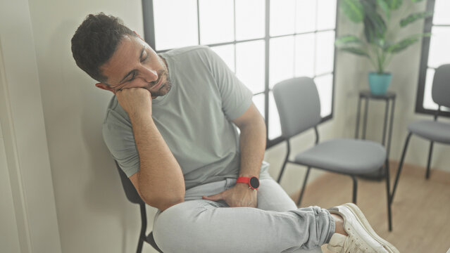 Man resting head on hand, sitting in chair by bright window, wearing gray tshirt, jeans and white sneakers in a clinic building waiting area; boredom fatigue.